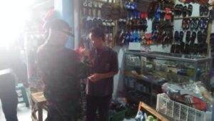 Seseorang dengan pakaian kamuflase dan penutup wajah berbicara dengan seorang penjaga toko di dalam toko kecil yang menjual sandal, sepatu, dan tas, dengan rak dan etalase kaca terlihat.