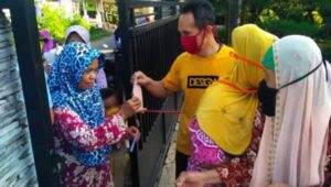 Seorang pria dengan kemeja kuning dan masker merah membagikan masker kepada para wanita berhijab yang berkumpul di gerbang luar ruangan pada siang hari. Para wanita menerima masker sambil berdiri bersama dalam kelompok kecil.